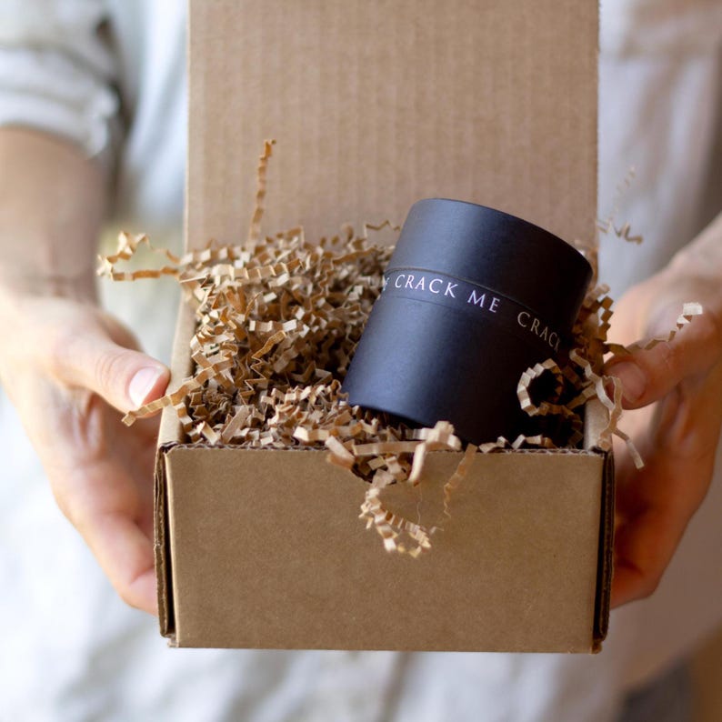 May include: A black cylindrical container with the text "CRACK ME" is nestled in shredded brown paper inside a cardboard box. The box is open, and the container is the focal point.