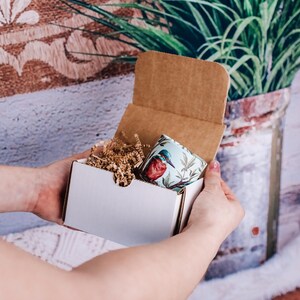 May include: A white cardboard box with a brown cardboard lid is open, revealing a small, colorful mug with a bird design. The mug is surrounded by brown packing peanuts.