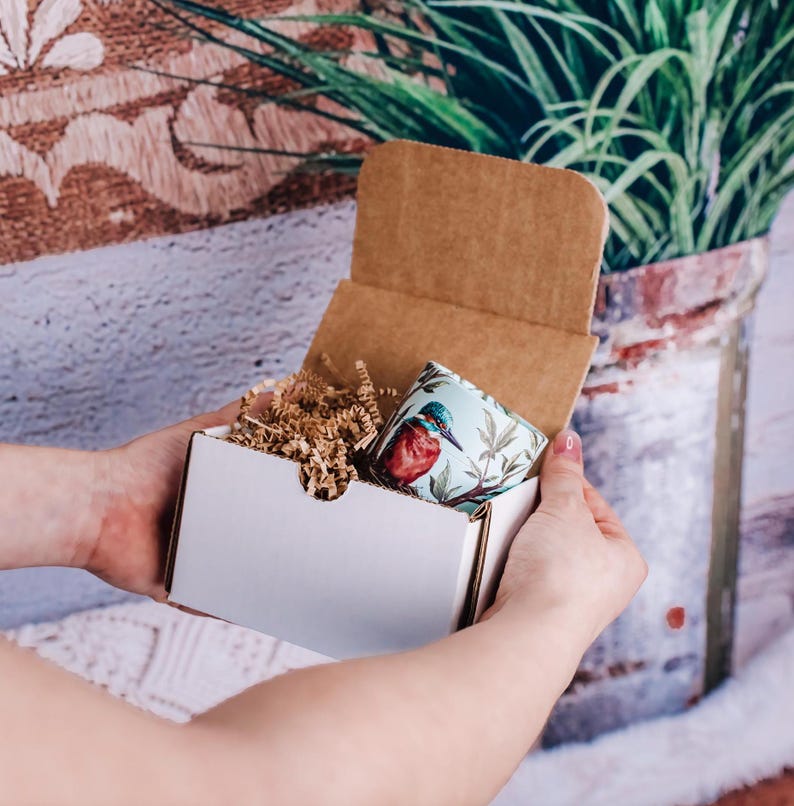 May include: A white cardboard box with a brown cardboard lid is open, revealing a small, colorful mug with a bird design. The mug is surrounded by brown packing peanuts.