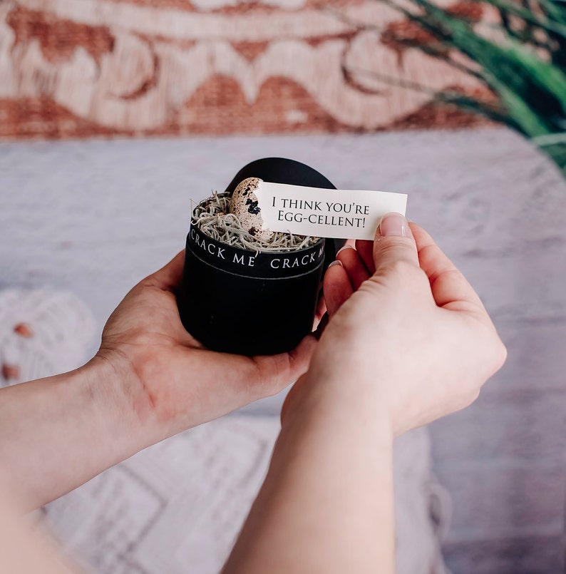 May include: A black velvet box with the words "CRACK ME" printed on the side, containing a speckled egg nestled in decorative filler. A small card reads "I THINK YOU'RE EGG-CELLENT!"