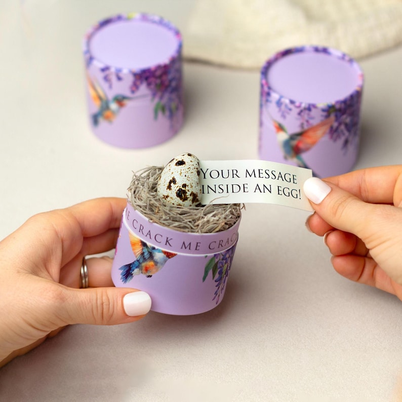 K&ouml;nnte beinhalten: Drei zylindrische Geschenkboxen in Lavendelfarbe mit Kolibri- und Blumendesigns. Eine Box ist ge&ouml;ffnet und zeigt ein Nest mit einem gesprenkelten Ei und einer Nachricht mit der Aufschrift &bdquo;YOUR MESSAGE INSIDE AN EGG!&ldquo;. Auf der Box steht auch &bdquo;CRACK ME CRACK&ldquo;.