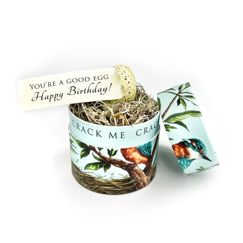 May include: A round gift box with a lid, featuring a bird and branch design in shades of blue, green, and orange. The box contains dried grass and a speckled egg. A tag reads "You're a good egg Happy Birthday!"