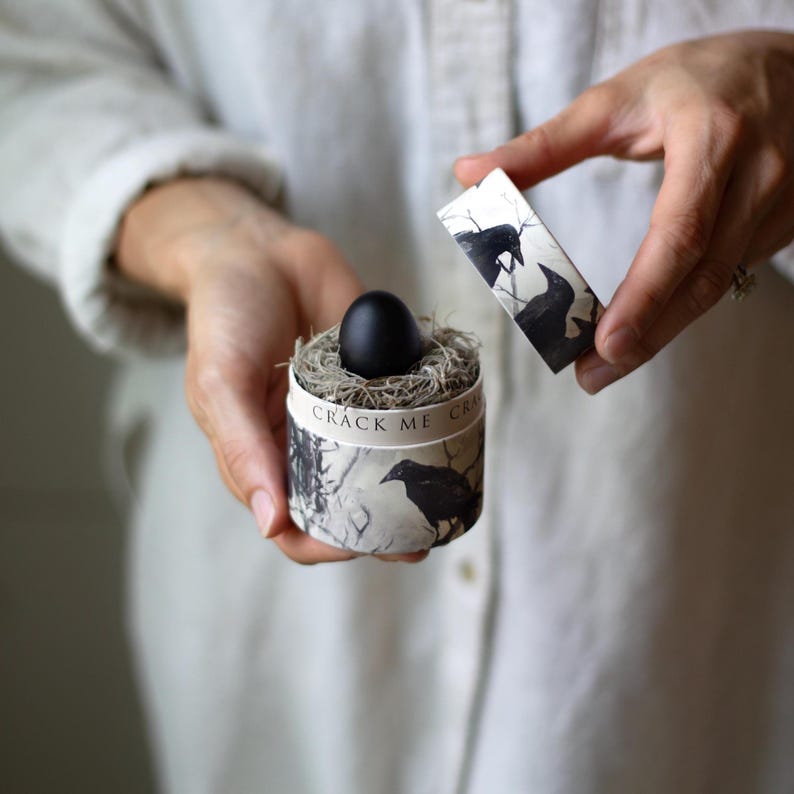 May include: A black egg rests in a nest of dried grass inside a round box. The box is decorated with a black and white crow illustration and the words "CRACK ME". The lid of the box is open, revealing the crow design.
