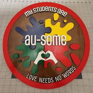 May include: A round wooden sign with a red border. The sign features a puzzle piece design in blue, yellow, green, and red, with the words "my students are au-some" and "love needs no words".