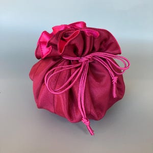 May include: A small, round, magenta-colored satin pouch with a drawstring closure. The pouch has a ruffled top edge and a contrasting bright pink lining. The drawstring is a matching bright pink cord, tied in a bow.