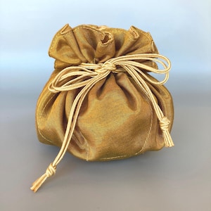 Könnte beinhalten: Ein kleiner, runder, goldgelber Beutel mit Kordelzug. Die Tasche besteht aus einem strukturierten Stoff und ist oben mit einem passenden Kordelzug gerafft. Der Kordelzug ist zu einer Schleife gebunden. Die Tasche steht auf einer grauen Oberfläche.