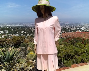 May include: A light pink skirt suit with a matching long-sleeved jacket and a pleated skirt. The mannequin is wearing a wide-brimmed green hat with a decorative band. The outfit is displayed outdoors with a city and blue sky in the background.