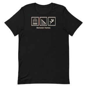 May include: A black short-sleeve t-shirt featuring a graphic of three film strip frames containing a window, a staircase, and a plant. Below the frames, the text "Between frames" is printed in a light beige color.
