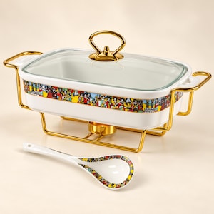 May include: A rectangular white ceramic food warmer with a glass lid and gold-toned metal stand. The dish features a colorful band of illustrated figures around the exterior. A matching white ceramic spoon with a patterned handle is included.