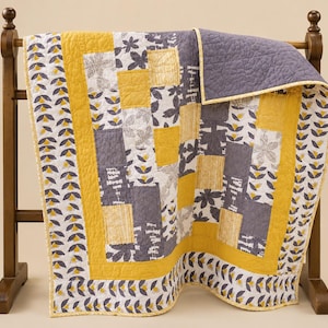 May include: A quilted blanket featuring a patchwork design of mustard yellow and charcoal gray fabric squares. The pattern includes floral motifs, vertical stripes, and solid blocks, finished with a mustard yellow border and a repeating leaf print.