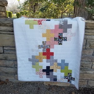 May include: A white quilt with a patchwork design featuring a variety of colours and patterns, including red, yellow, green, blue, and grey. The quilt is made up of squares with a plus sign design in the centre.