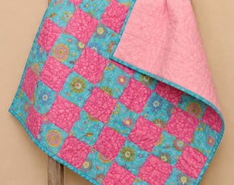 Puede incluir: Una manta acolchada con un patrón de tablero de ajedrez de cuadrados de color rosa intenso sólido y cuadrados de color azul claro con un estampado floral multicolor. La manta tiene un reverso de color rosa claro sólido y un borde de tela azul claro.
