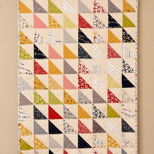 Peut inclure: Une courtepointe rectangulaire composée d'une grille de triangles rectangles de différentes couleurs, notamment rouge, jaune, vert, bleu marine et gris. Les tissus présentent des motifs variés comme des pois, des croix et du texte sur fond crème.