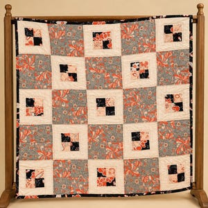 May include: A square quilt featuring a repeating patchwork pattern of cream-colored squares alternating with blocks of gray fabric printed with orange and white floral motifs, interspersed with smaller four-patch squares of black and peach fabric.