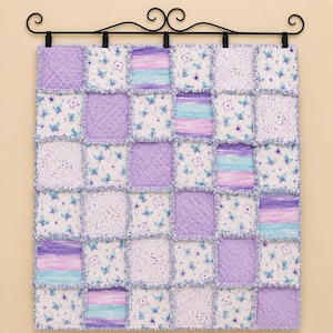 Könnte beinhalten: Eine quadratische Patchwork-Decke bestehend aus Quadraten aus lavendelfarbenem Noppenstoff, weißem Stoff mit blauen Schmetterlingsmustern und horizontal gestreiftem Stoff in Lila und Türkis, aufgehängt an einer schwarzen Metallhalterung.