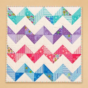 May include: A square quilt featuring a chevron pattern composed of alternating white fabric and colorful triangular patches. The patches display various floral, geometric, and dotted prints in shades of turquoise, purple, pink, and blue, finished with a striped border.