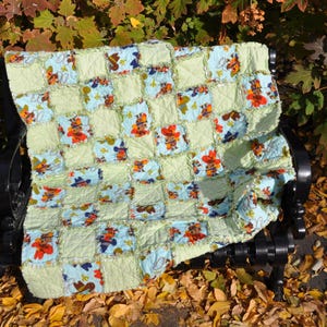 May include: A handmade patchwork quilt with a cowboy theme. The quilt features a repeating pattern of light blue squares with cowboy illustrations, alternating with light green squares. The quilt is draped over a black bench, with autumn leaves in the foreground.