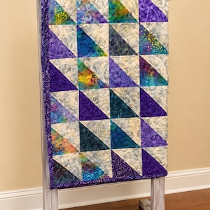 May include: A quilt featuring a half-square triangle pattern composed of various fabric squares. The design includes shades of purple, teal, blue, and yellow, contrasted with light gray marbled fabric. The quilt has a dark purple speckled border.