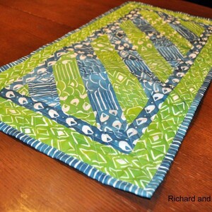 Peut inclure: Chemin de table matelassé rectangulaire avec un design vibrant. Le chemin de table présente un patchwork de tissus verts et bleus avec des accents blancs. Les bords sont finis avec une bordure rayée bleue et blanche. L'esthétique générale est faite à la main et décorative.