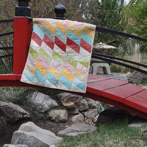 Puede incluir: Una colcha de retazos colorida con un patrón geométrico de triángulos en tonos azules, amarillos, rosas y rojos está drapeada sobre un puente de madera rojo.