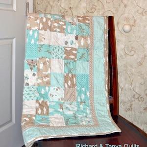 May include: A light blue and brown baby quilt with a patchwork design featuring clouds, trees, and a castle. The quilt has a white border and is folded on a wooden chair. The text "Richard & Tanya Quilts" is visible in the bottom right corner of the image.