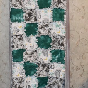 Puede incluir: Una colcha de bebé hecha a mano con un diseño de patchwork. La colcha presenta cuadrados alternos de color verde oscuro, tie-dye gris y tela blanca con ilustraciones de animales. La colcha se exhibe en una escalera de madera.