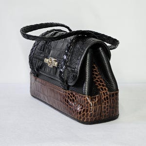 May include: A black and brown handbag with a crocodile skin pattern. The top portion is black with a braided handle and a decorative clasp. The bottom section is brown, and the bag has a rectangular shape.