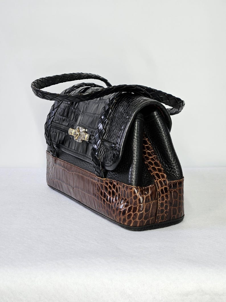 May include: A black and brown handbag with a crocodile skin pattern. The top portion is black with a braided handle and a decorative clasp. The bottom section is brown, and the bag has a rectangular shape.