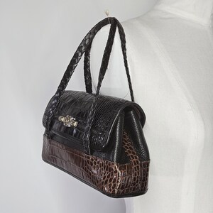 May include: A black and brown handbag with a crocodile pattern. The bag has a braided handle and a decorative clasp. The bottom of the bag is brown, while the top is black. The bag is hanging from a hook.