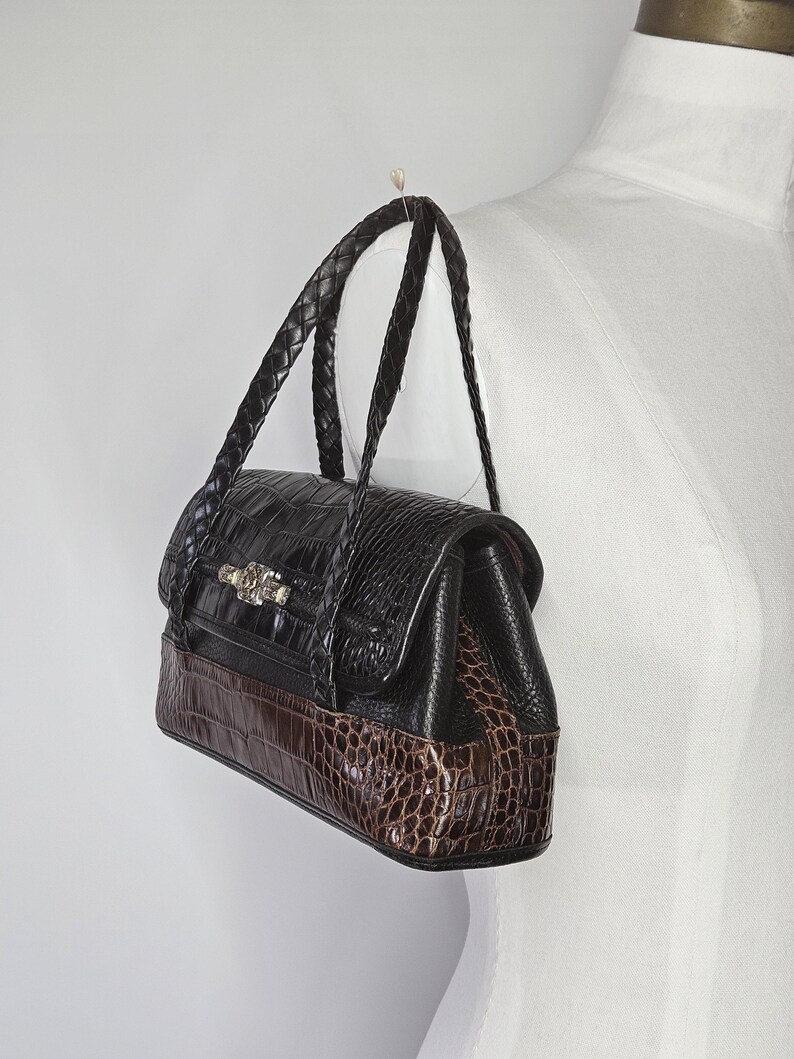 May include: A black and brown handbag with a crocodile pattern. The bag has a braided handle and a decorative clasp. The bottom of the bag is brown, while the top is black. The bag is hanging from a hook.