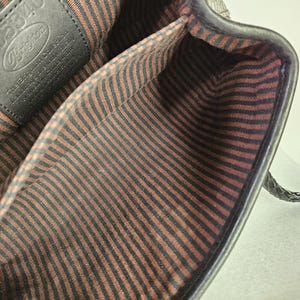 May include: Black leather handbag with a braided handle. The interior features a brown and black striped fabric lining. A black leather tag with the Brighton logo is visible inside the bag.