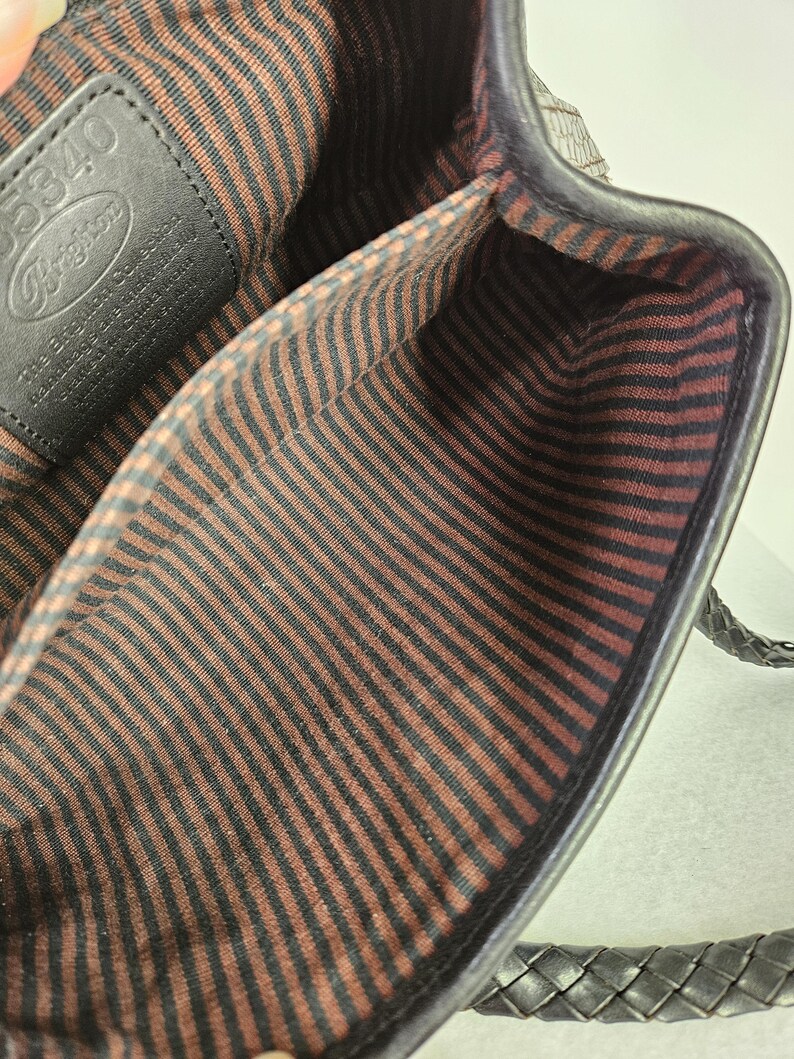 May include: Black leather handbag with a braided handle. The interior features a brown and black striped fabric lining. A black leather tag with the Brighton logo is visible inside the bag.