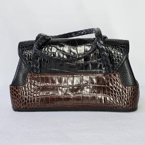 May include: A two-tone handbag with a crocodile skin pattern. The top portion is black, and the bottom is brown. The bag has a braided handle and a flap closure. The bag is rectangular and sits on a white surface.