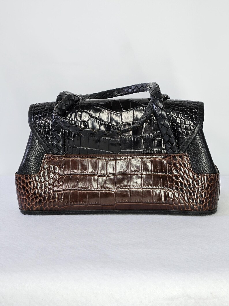 May include: A two-tone handbag with a crocodile skin pattern. The top portion is black, and the bottom is brown. The bag has a braided handle and a flap closure. The bag is rectangular and sits on a white surface.