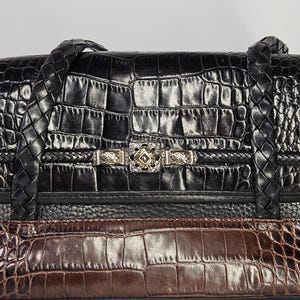 May include: Black and brown leather handbag with a crocodile skin pattern. The bag features braided handles and a silver-toned clasp with decorative details. The bag is divided into sections of black and brown leather.