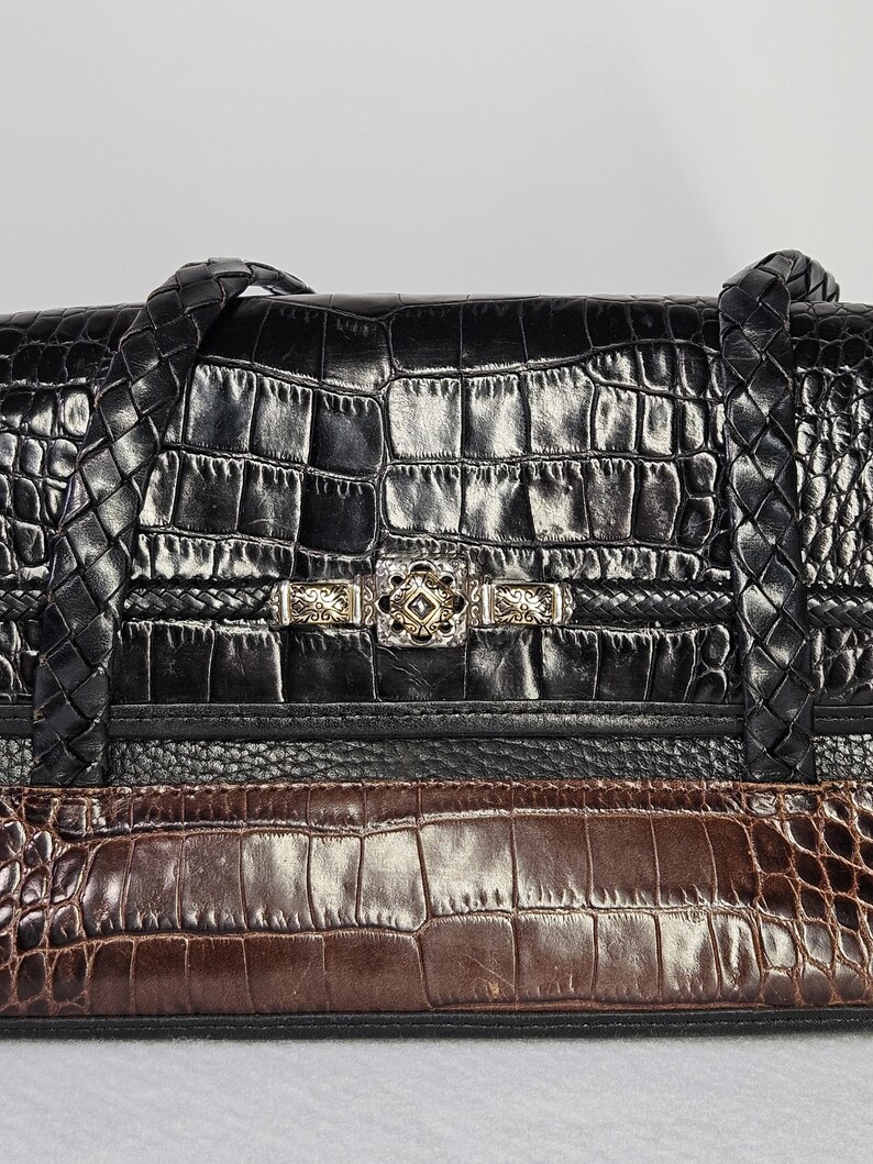 May include: Black and brown leather handbag with a crocodile skin pattern. The bag features braided handles and a silver-toned clasp with decorative details. The bag is divided into sections of black and brown leather.