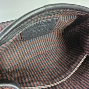 May include: Interior view of a black leather handbag. The inside features a brown and black striped fabric lining. A small black leather tag with embossed text is visible. The bag's edges are smooth and dark.