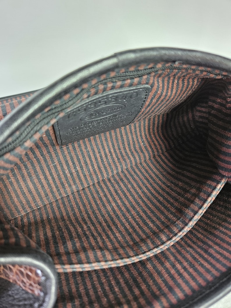 May include: Interior view of a black leather handbag. The inside features a brown and black striped fabric lining. A small black leather tag with embossed text is visible. The bag's edges are smooth and dark.