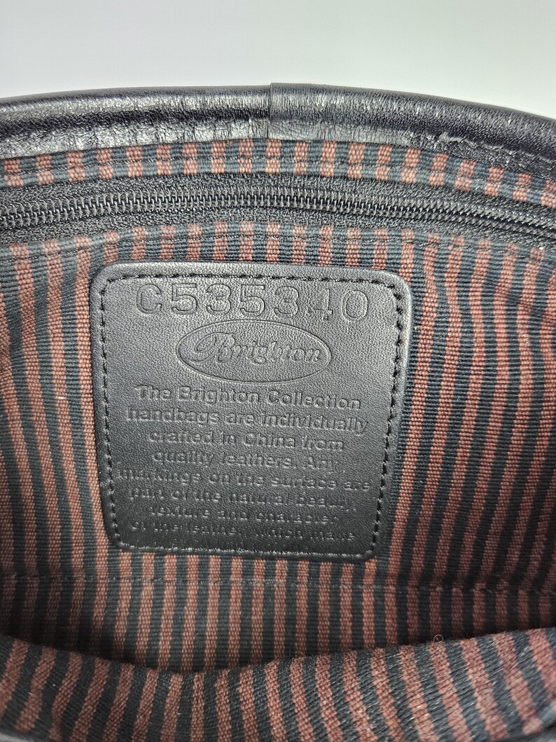 May include: Interior view of a black leather handbag. The lining features brown and black vertical stripes. A black leather tag displays the Brighton logo and the text "The Brighton Collection handbags are individually crafted in China from quality leathers."
