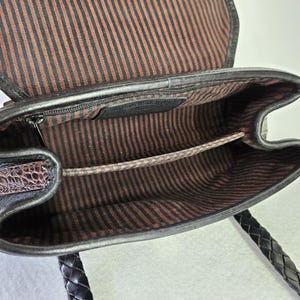 May include: Open view of a black leather handbag with a braided handle. The interior features a brown and black striped fabric lining. A small zippered pocket is visible inside the bag. The bag's flap is open, revealing the interior.