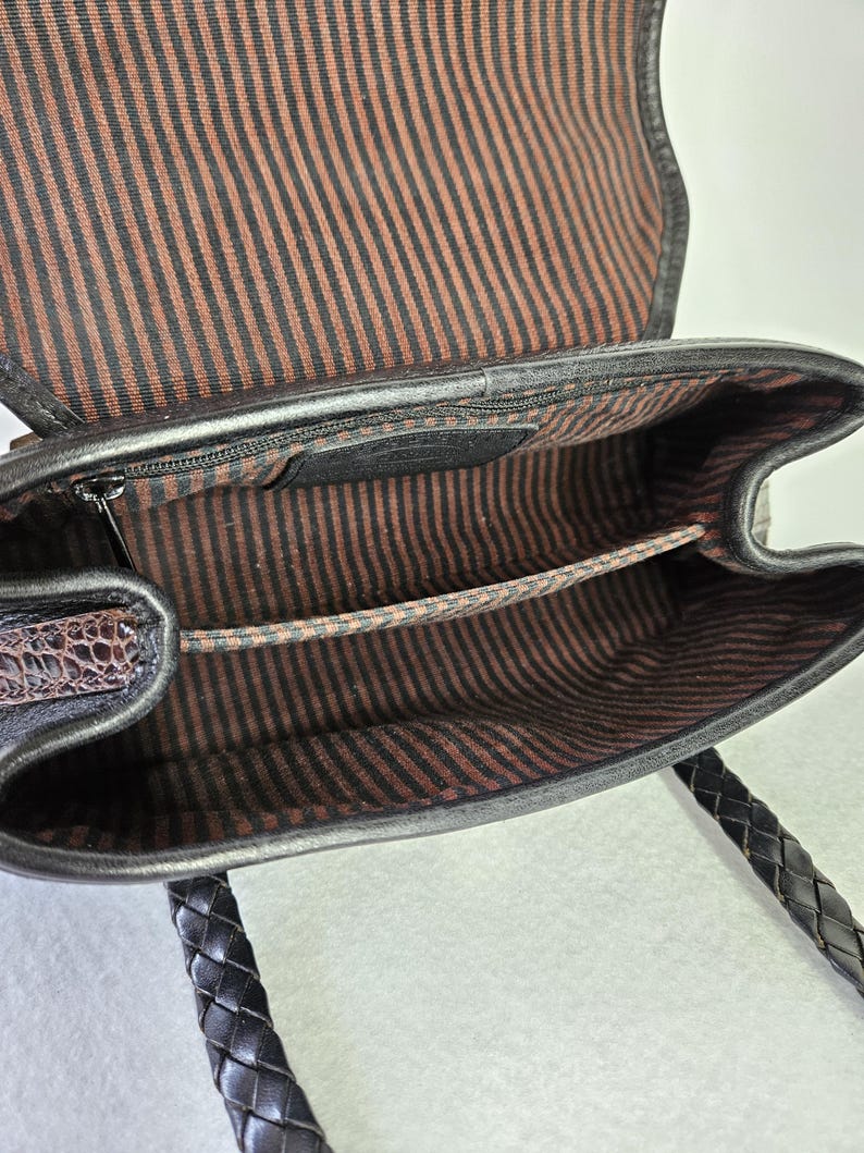 May include: Open view of a black leather handbag with a braided handle. The interior features a brown and black striped fabric lining. A small zippered pocket is visible inside the bag. The bag's flap is open, revealing the interior.