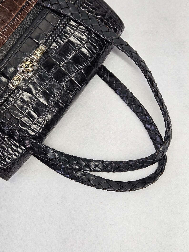 May include: Black leather handbag with a crocodile-skin texture. The bag features braided handles and a decorative silver clasp with a square, green stone. The top of the bag is brown leather.