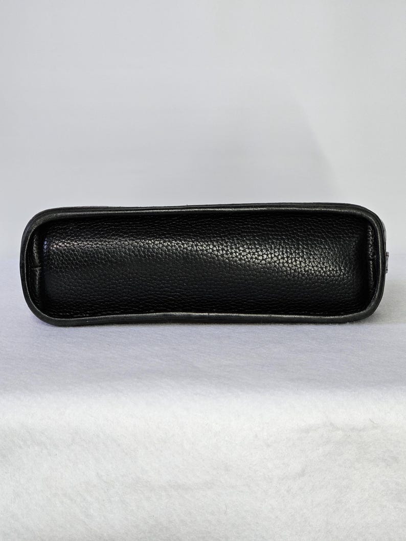 May include: A black leather handbag with a textured surface. The bag has a rectangular shape with rounded edges and a smooth, dark border. The bag is designed to be carried by hand or over the shoulder.