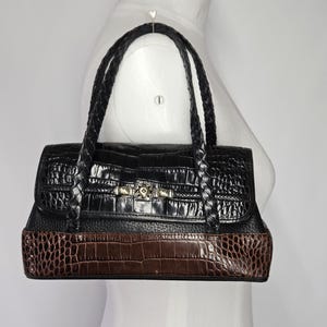 May include: Black and brown handbag with a crocodile-textured design. The top portion is black, while the bottom is brown. The bag has braided handles and a gold-colored clasp. The bag is displayed on a white mannequin.