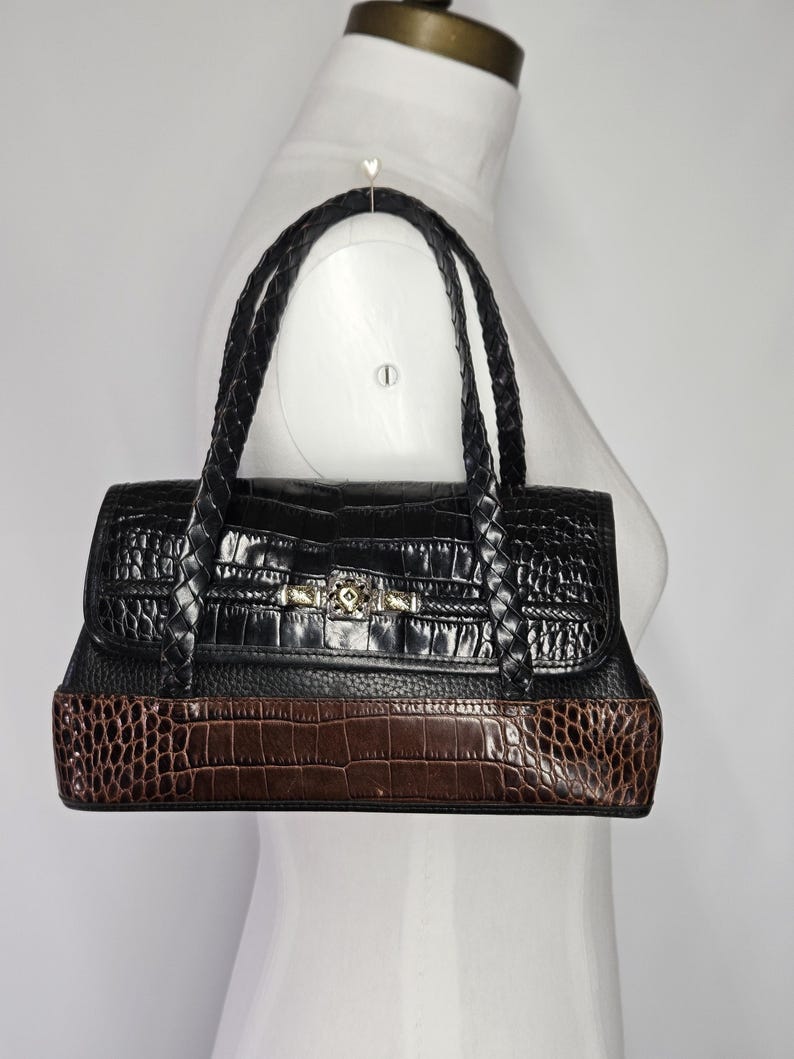 May include: Black and brown handbag with a crocodile-textured design. The top portion is black, while the bottom is brown. The bag has braided handles and a gold-colored clasp. The bag is displayed on a white mannequin.