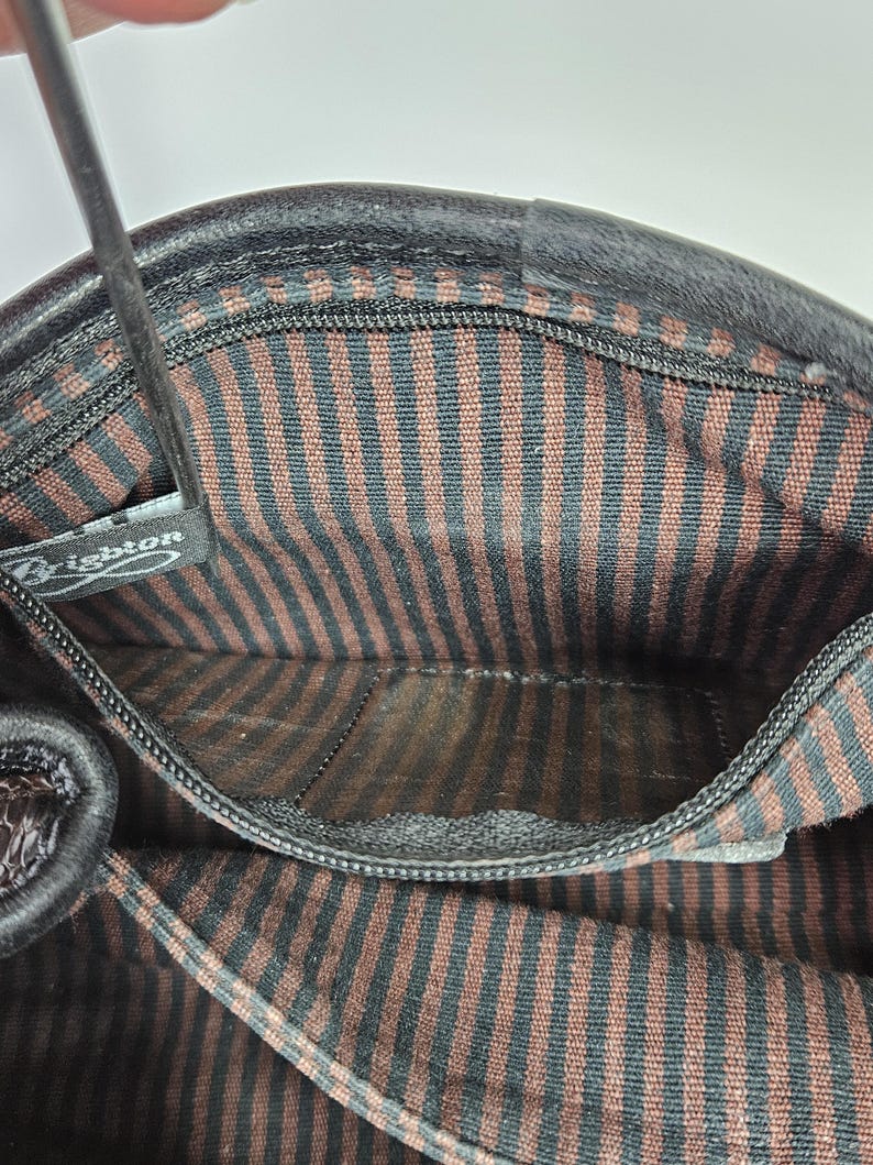 May include: Interior view of a black leather handbag. The lining features brown and black vertical stripes. A small zippered pocket is visible. The brand name "Brighton" is visible on a tag.