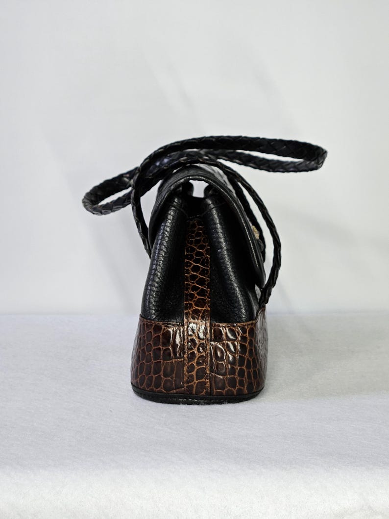 May include: A black and brown handbag with a braided handle. The top portion is black, while the bottom is brown with a crocodile pattern. The bag has a flap closure and a gold-colored clasp.