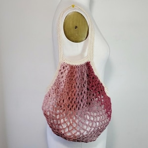 May include: A handmade crochet tote bag in shades of pink and burgundy. The bag features a net-like design with a cream-colored strap and trim. The bag is displayed on a mannequin.