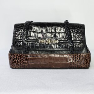 May include: A black and brown handbag with a crocodile skin pattern. The top portion is black, and the bottom is brown. The bag has a braided handle and a decorative clasp. The bag is rectangular and sits on a white surface.