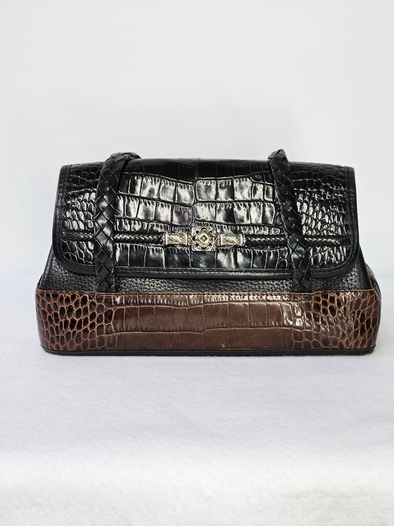 May include: A black and brown handbag with a crocodile skin pattern. The top portion is black, and the bottom is brown. The bag has a braided handle and a decorative clasp. The bag is rectangular and sits on a white surface.